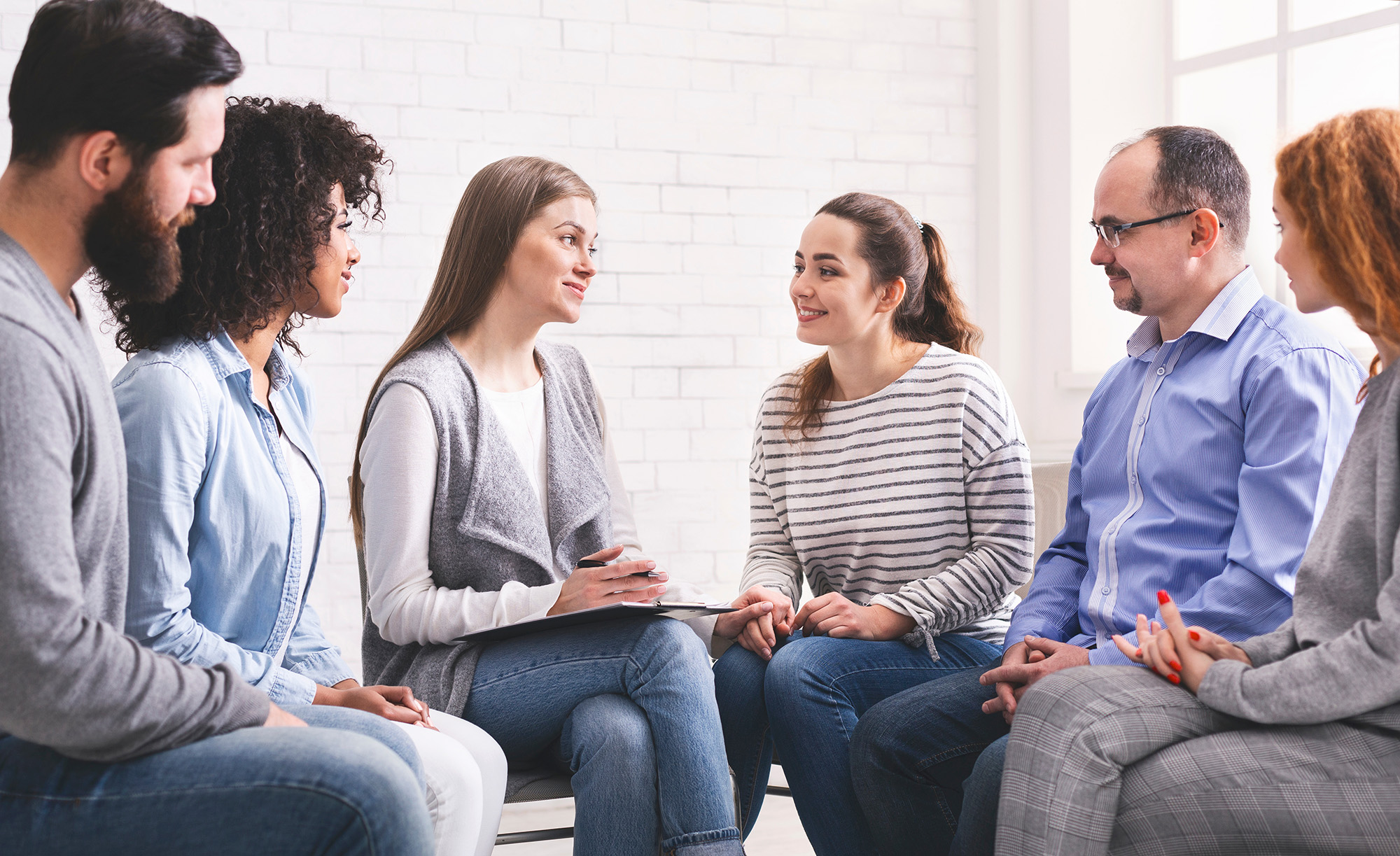 A group of six individuals, including one man and five women, gathered in a brightly lit room with a brick wall, engaged in conversation around a table.