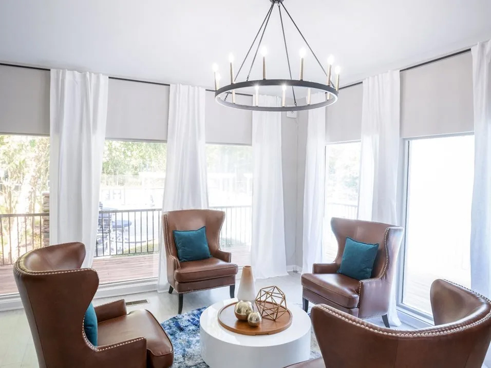 The image shows an interior space, possibly a living room or den, with two leather armchairs facing each other around a small round table. There is a large chandelier hanging from the ceiling and white curtains framing a window that lets in natural light. The room has a modern aesthetic with a neutral color palette and a combination of brown leather furniture and blue accents.