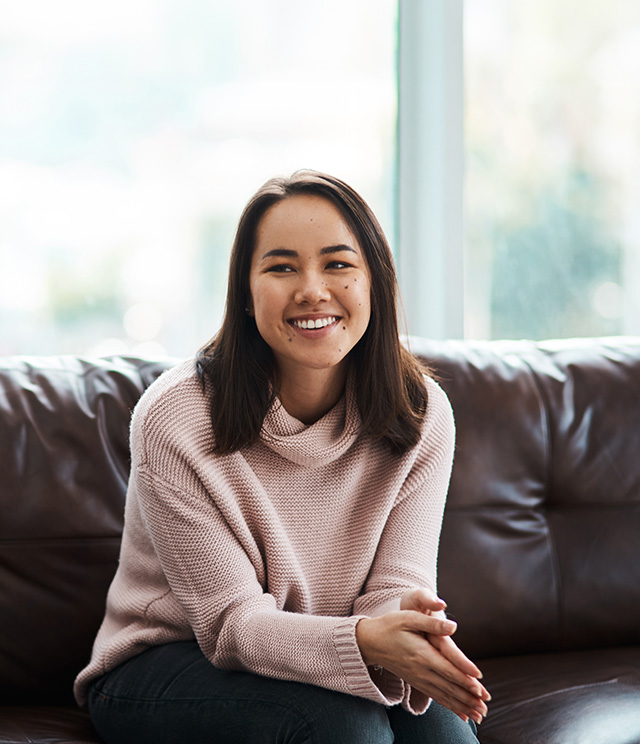 A woman sitting on a couch with her hands clasped in front of her, smiling and looking directly at the camera.